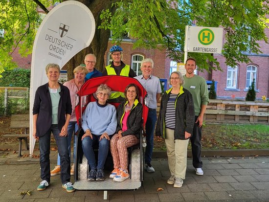 Zwei Personen sitzen im roten Rollstuhl, umgeben von sieben weiteren Personen im Freien. Links ist ein weißes Banner des Deutschen Ordnungsdienstes, rechts ein Bushaltestellenschild zu sehen. Im Hintergrund sind Bäume und ein Backsteingebäude erkennbar.