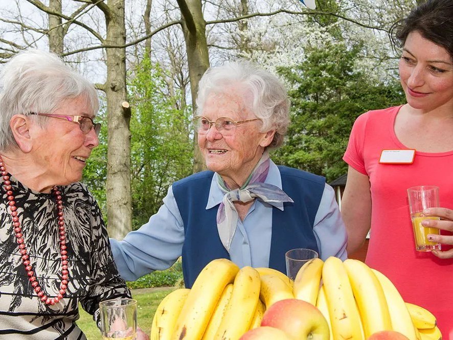 Zwei Bewohnerinnen im Garten: vor ihnen eine Obstschale mit Äpfeln und Bananen - eine Pflegerin steht mit einem Glas O-Saft daneben