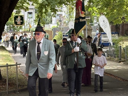 Männer in traditioneller grüner Tracht marschieren in einer Prozession auf einem Weg. Im Vordergrund führt ein Mann im grauen Jackett und Hut die Gruppe an, während ein anderer eine große Fahne mit einer religiösen Darstellung trägt. Im Hintergrund sind weitere Teilnehmer und eine Blaskapelle zu erkennen.