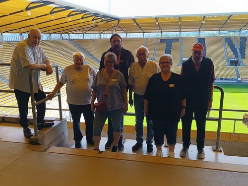 Sieben Personen posieren für ein Gruppenfoto auf einer Tribüne in einem Fußballstadion. Im Hintergrund sind gelbe Sitzreihen und der grüne Rasen zu erkennen.