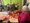 Senioren sitzen in einem Raum mit grünen Wänden. Im Vordergrund bereitet eine Frau in einem rosa Pullover einen Erdbeerkuchen zu, während ein Mann daneben Früchte schneidet. Im Hintergrund essen weitere Senioren an Tischen.