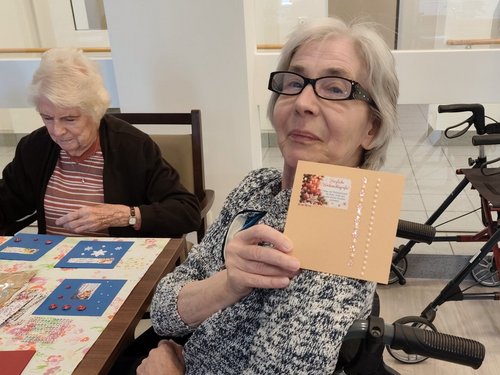 Eine ältere Frau im Rollstuhl präsentiert eine selbstgebastelte Weihnachtskarte mit der Aufschrift „Herzliche Weihnachtsgrüße". Im Hintergrund sitzt eine weitere Dame an einem Tisch und bastelt.