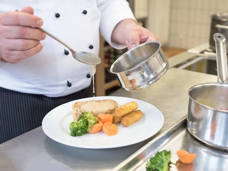 Nahaufnahme in der Küche: ein Koch gibt Soße auf einen Teller mit Fleisch und Gemüse