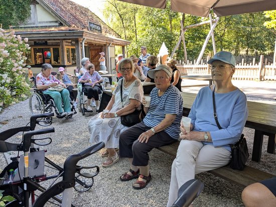 Eine Gruppe älterer Menschen sitzt entspannt auf Holzbänken und in Rollstühlen im Freien. Im Hintergrund ist ein kleines Gebäude zu sehen, umgeben von Bäumen. Die Szene zeigt eine friedliche Pause.