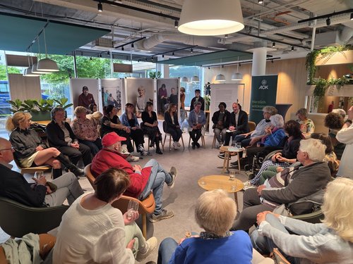 Eine Gruppe von Menschen sitzt in einem hellen, modernen Raum in einem großen Kreis. Im Hintergrund sind große Fenster, Pflanzen und Ausstellungstafeln zu sehen. Die Personen scheinen an einer Diskussion oder einem geselligen Treffen teilzunehmen.