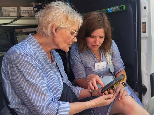 Zwei Frauen sitzen neben einem weißen Van mit dem Logo „Digitaler Engel". Eine jüngere Frau erklärt einer älteren Dame die Nutzung eines Tablets, auf das sie zeigt.
